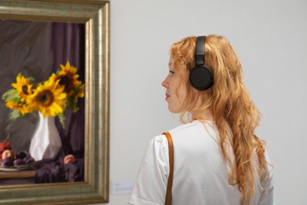 Mujer observando una pintura de girasoles en un museo, utilizando auriculares para enriquecer su experiencia al analizar una obra de arte.